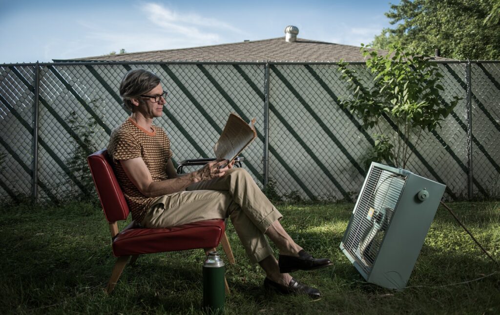 Dylan Hicks sits reading is his sunny backyard, a box fan pointed at him.